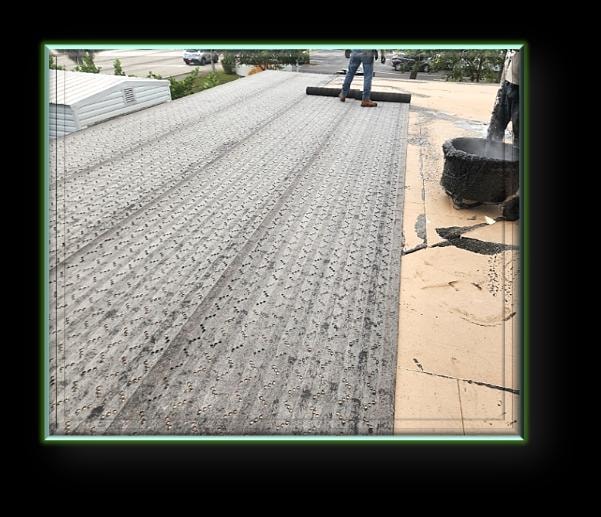 Worker rolling out modified bitumen base sheet across commercial flat roof deck with hot asphalt – Artisan Roof Specialists